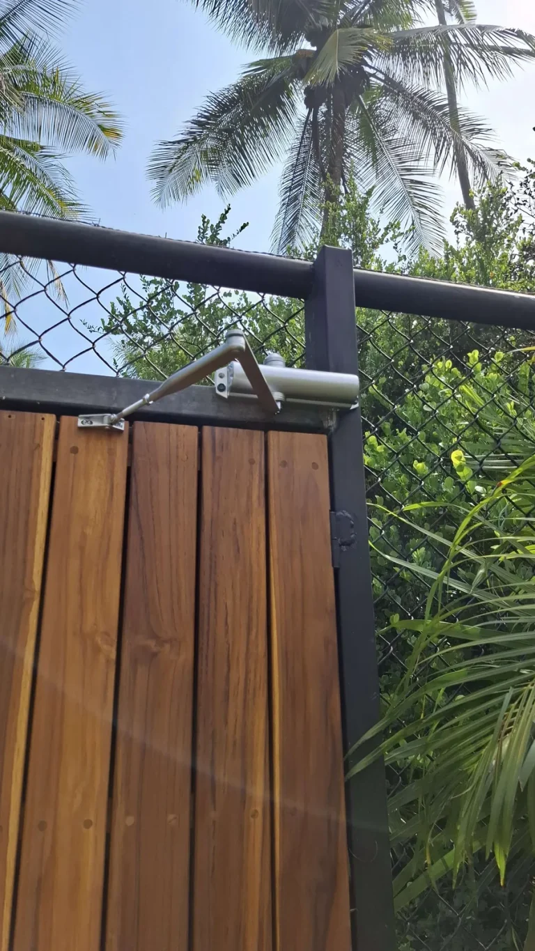Brazo cierrapuertas hidráulico plateado instalado en marco metálico de portón con lamas de madera.