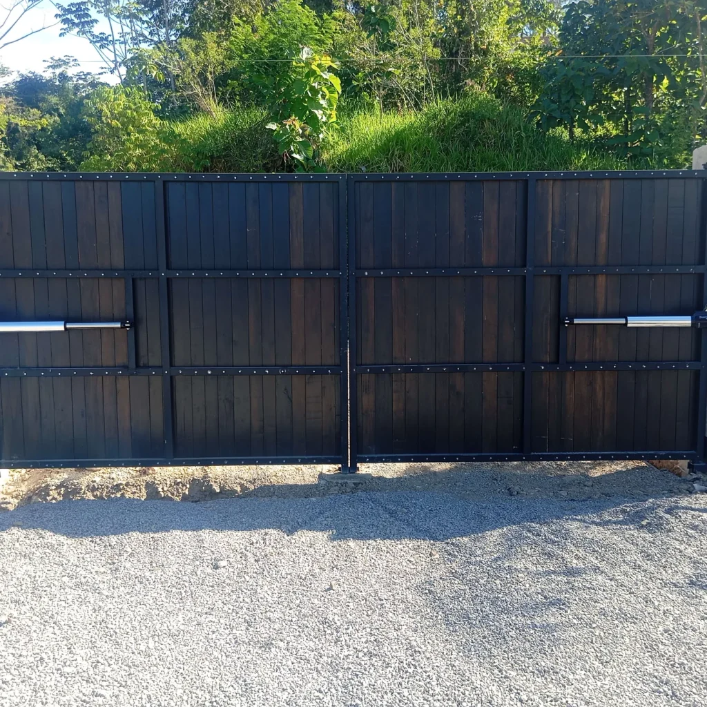 Portón eléctrico de dos hojas de madera oscura con marco negro y actuadores lineales montados en pilares de concreto
