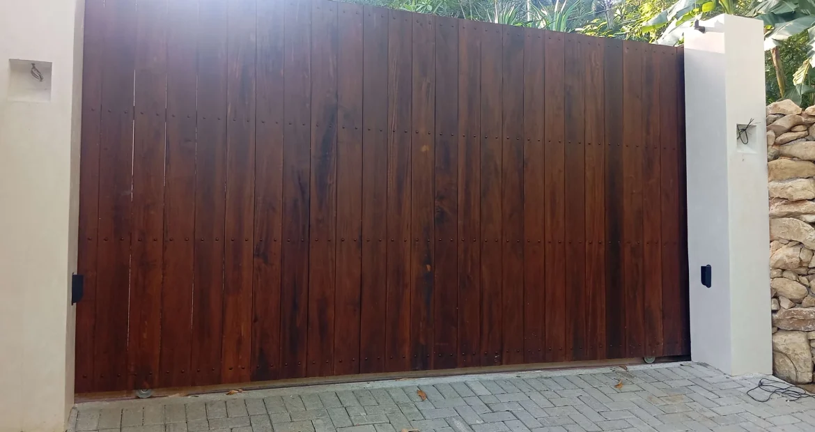 Portón eléctrico corredizo de madera oscura entre pilares blancos con entrada de adoquines y fondo tropical