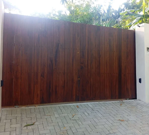 Portón eléctrico corredizo de madera oscura entre pilares blancos con entrada de adoquines y fondo tropical